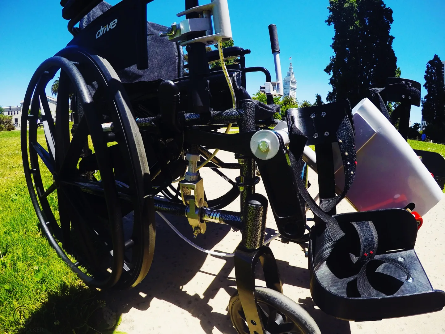 Close-up of LegMaker prototype mounting hardware, pedals and foot straps on a manual wheelchair outdoors