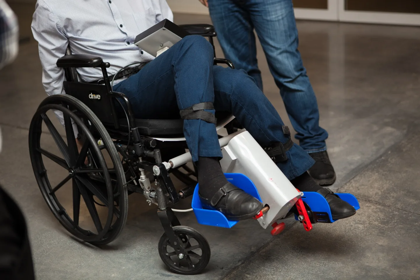 Person seated in a manual wheelchair with feet secured in the blue pedals of the LegMaker development prototype